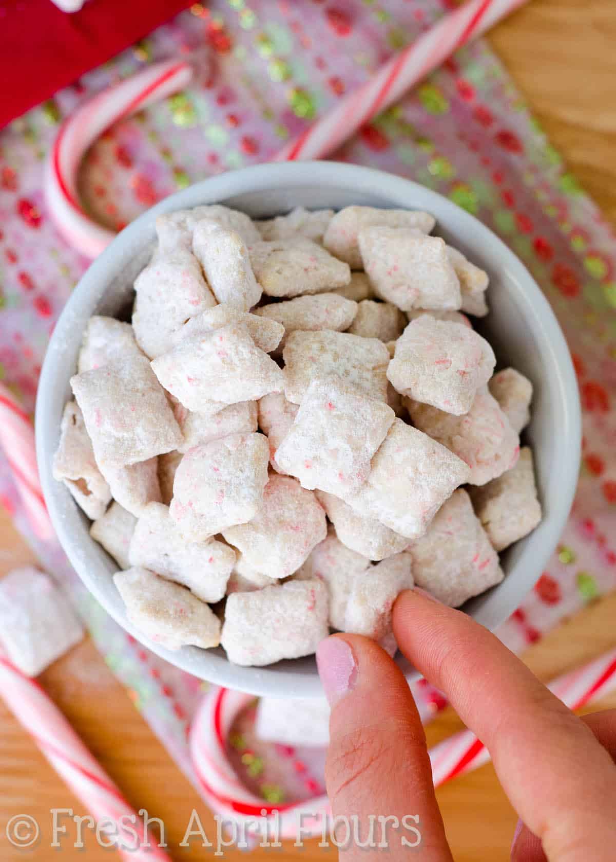 A bowl of candy cane puppy chow with fingers reaching in to taking a piece.
