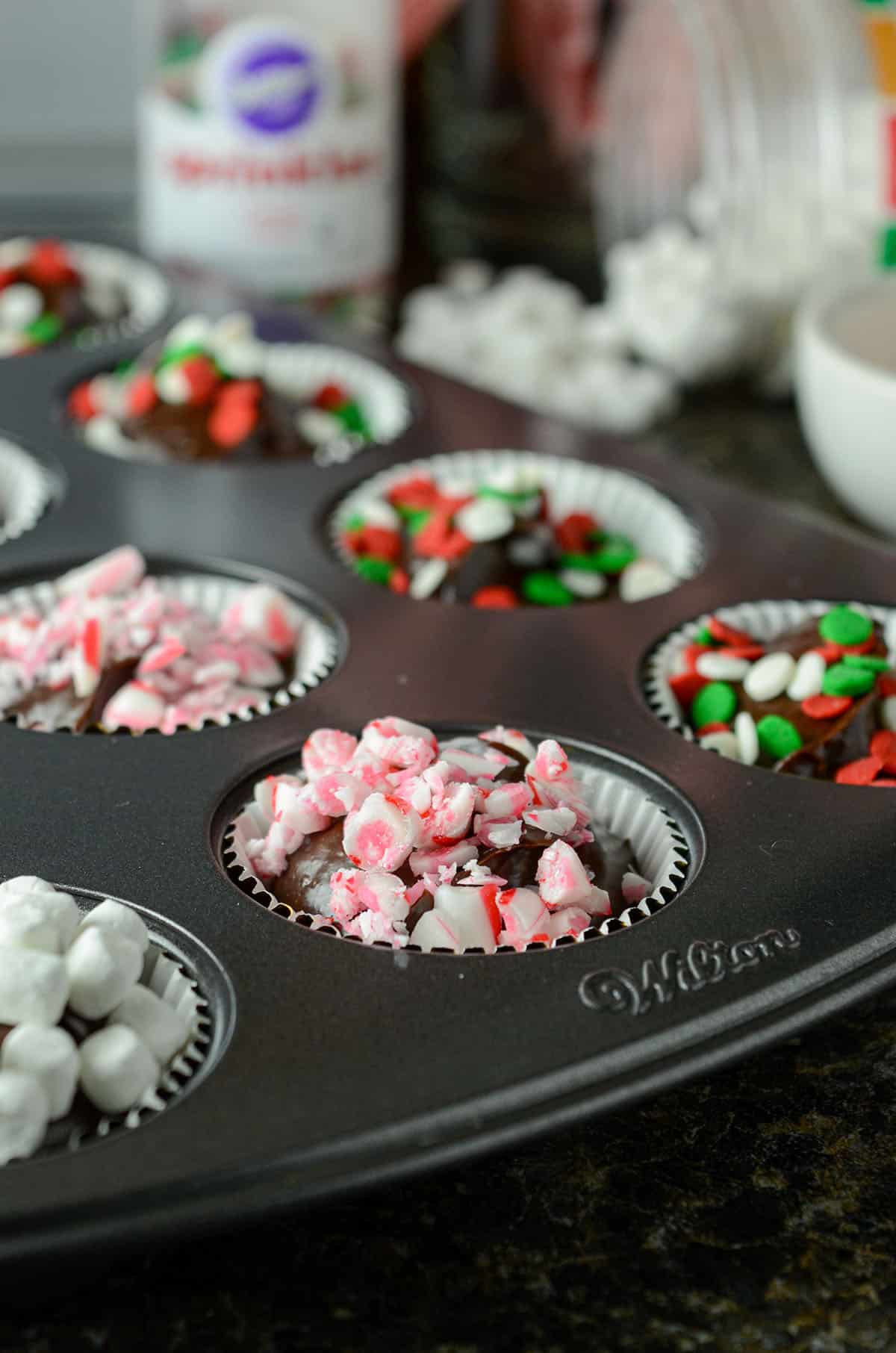 hot chocolate bombs sitting in mini-cupcake pan-- some have marshmallows, some have crushed candy canes, and some have christmas sprinkles