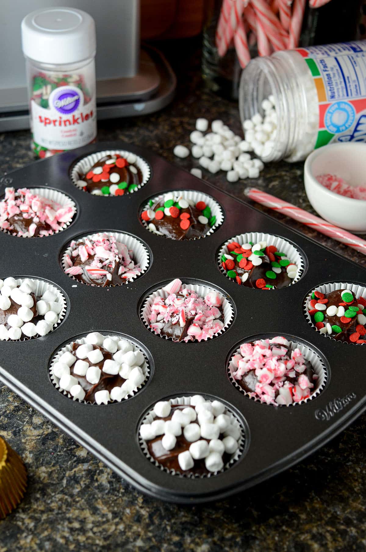 hot chocolate bombs sitting in mini-cupcake pan-- some have marshmallows, some have crushed candy canes, and some have christmas sprinkles