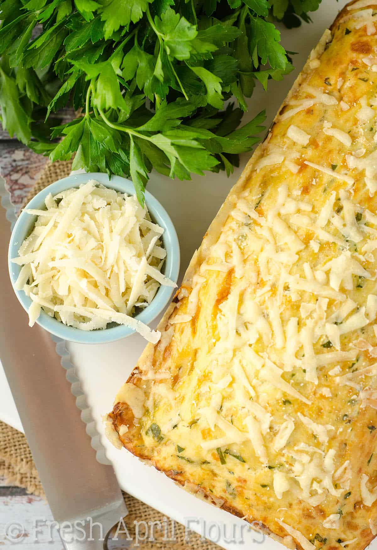 A loaf of parmesan parsley bread on a cutting board with a bowl of shredded cheese and a bunch of parsley.