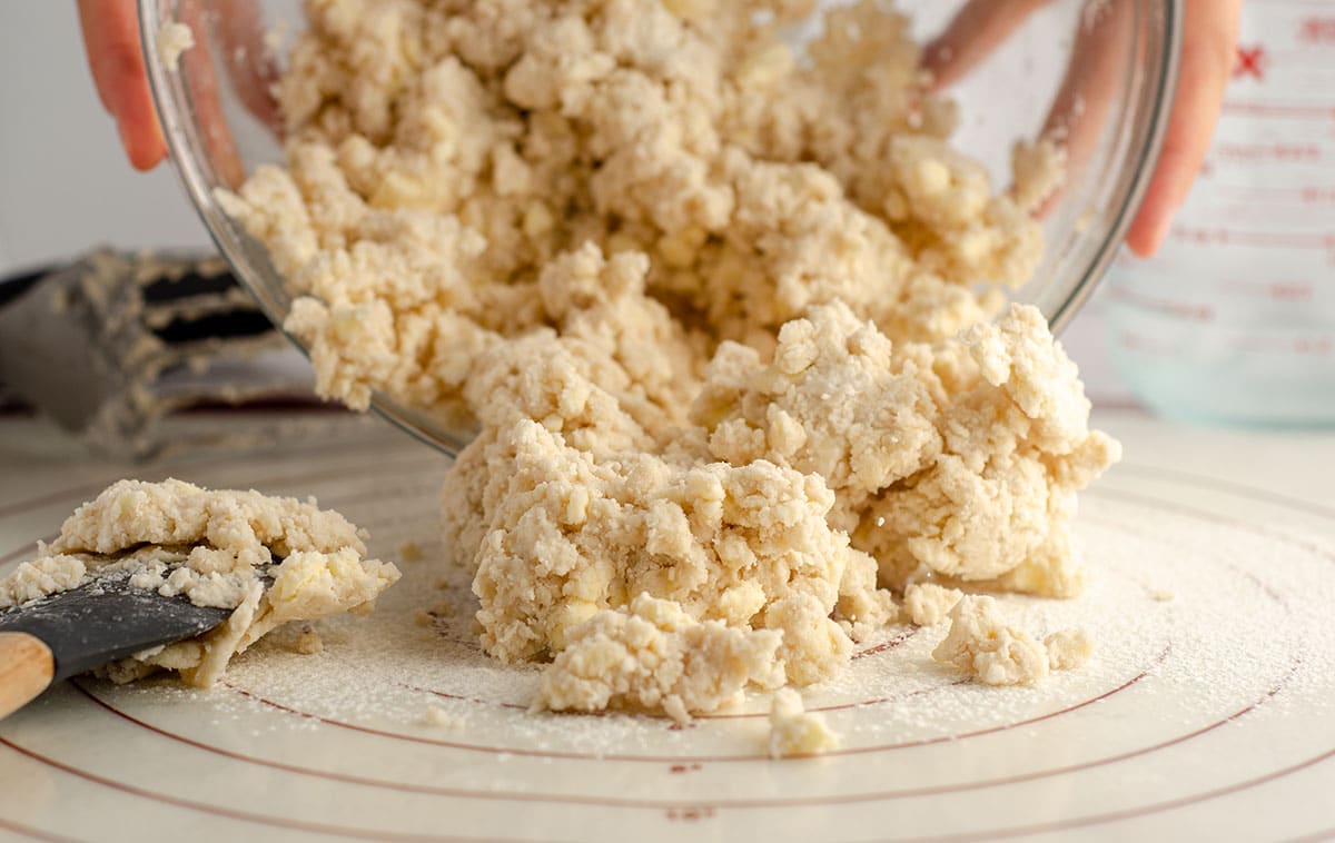 hands dumping crumbly pie dough out onto a pastry mat