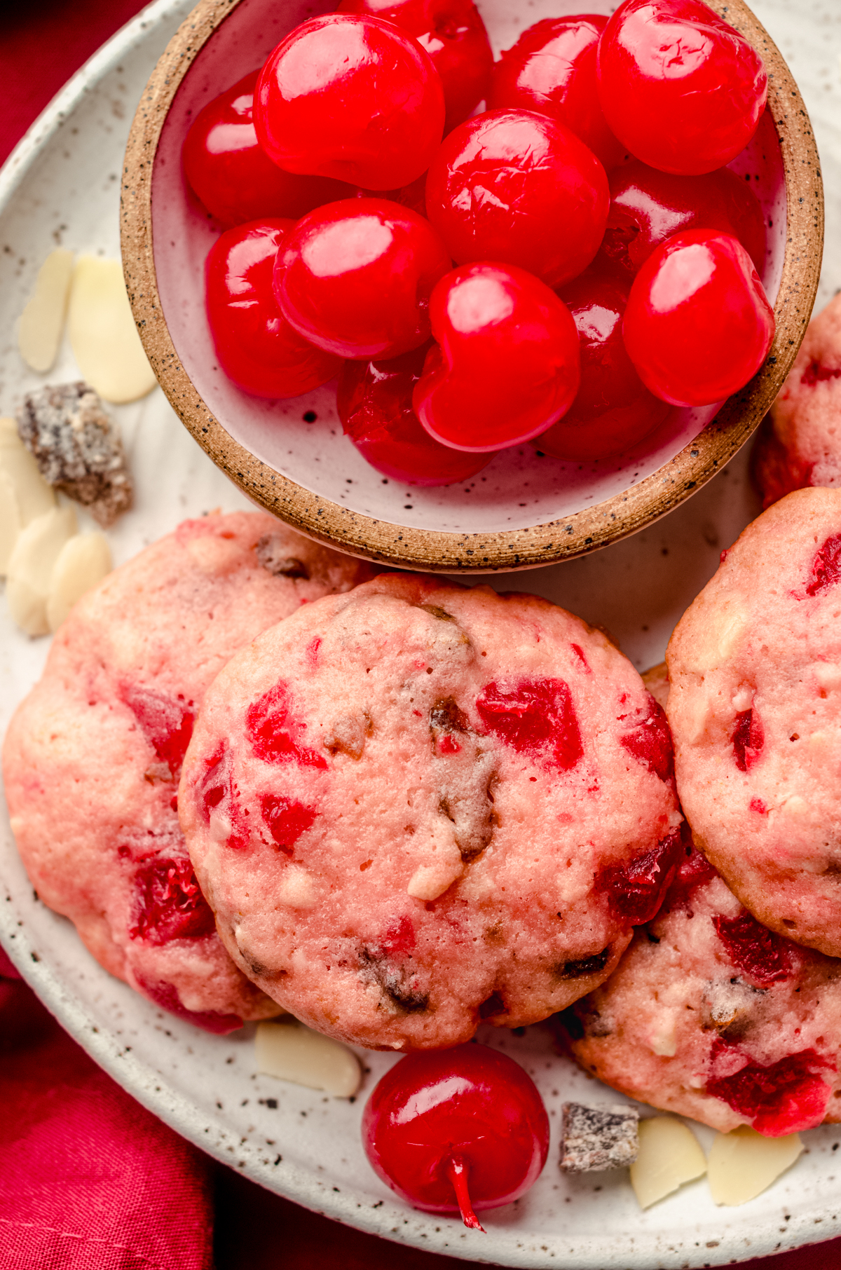Aerial photo of cherry almond date cookies on a plate with a small bowl of maraschino cherries.
