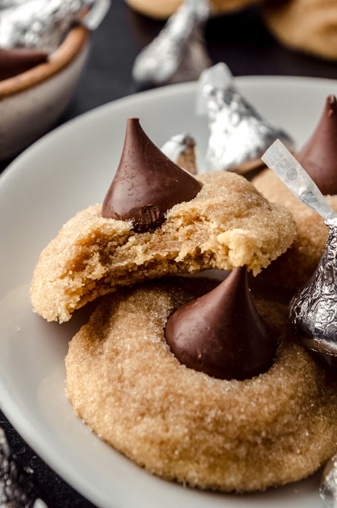 A plate of peanut butter blossoms and one has a bite taken out of it and there are Hershey's Kisses around it.