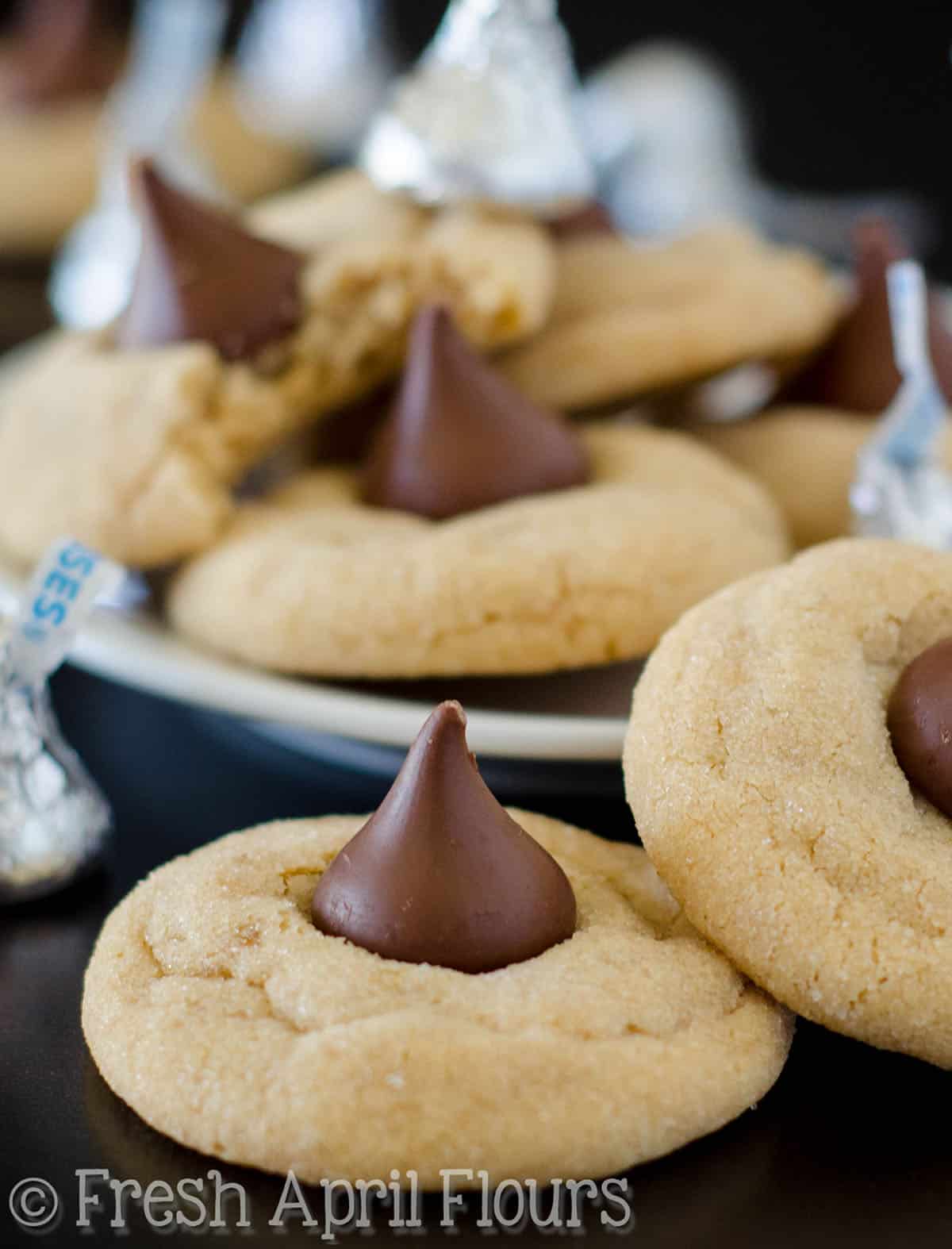 Peanut butter blossom cookies on a surface with a plate full of them in the background.