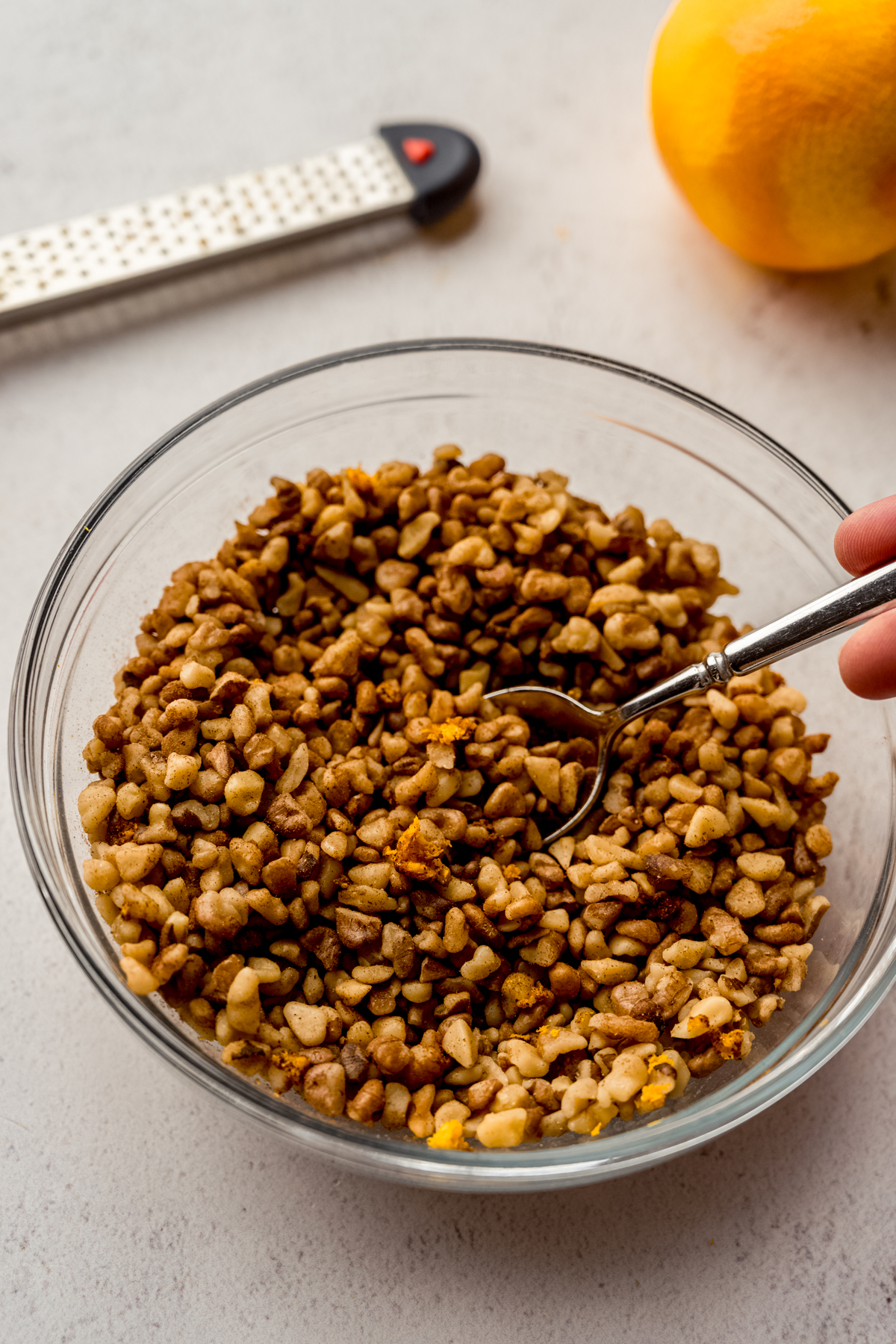 A bowl of walnuts with spices and orange zest in it that someone is stirring with a spoon.