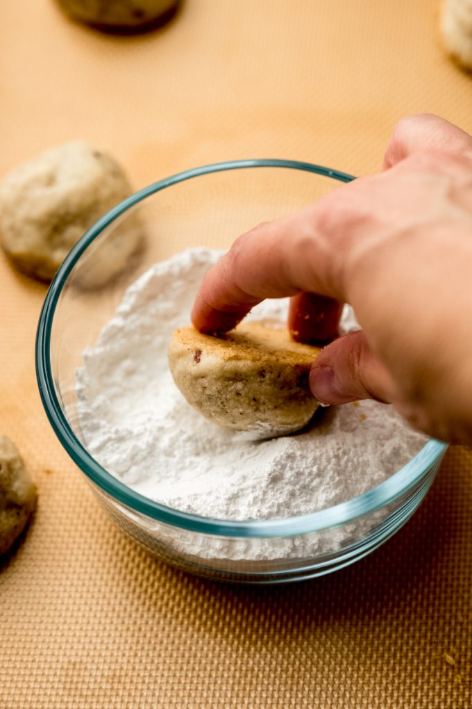Someone is rolling a snowball cookie in a bowl of powdered sugar.