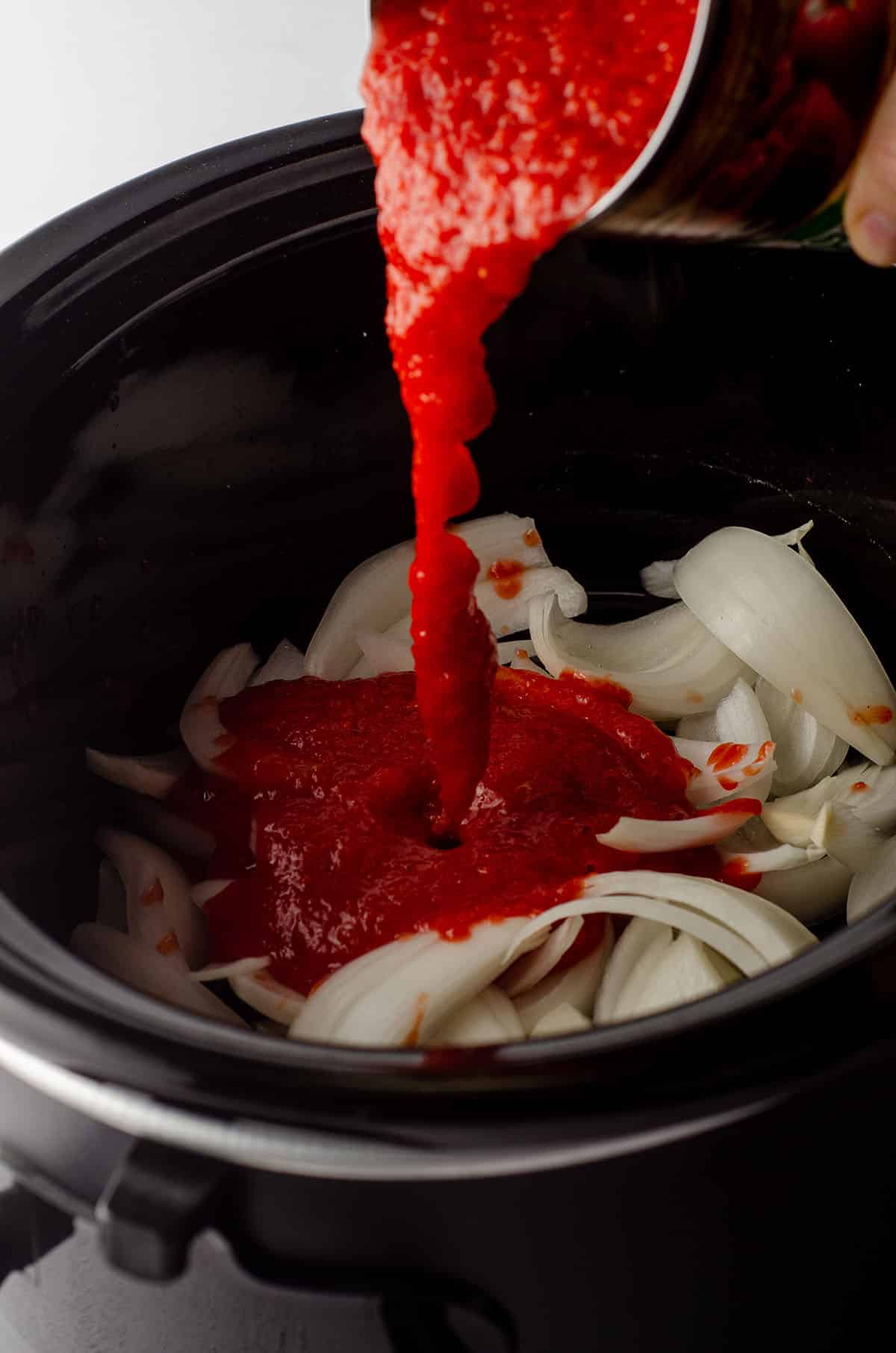 slow cooker filled with sliced onions and a can of crushed tomatoes being poured on top