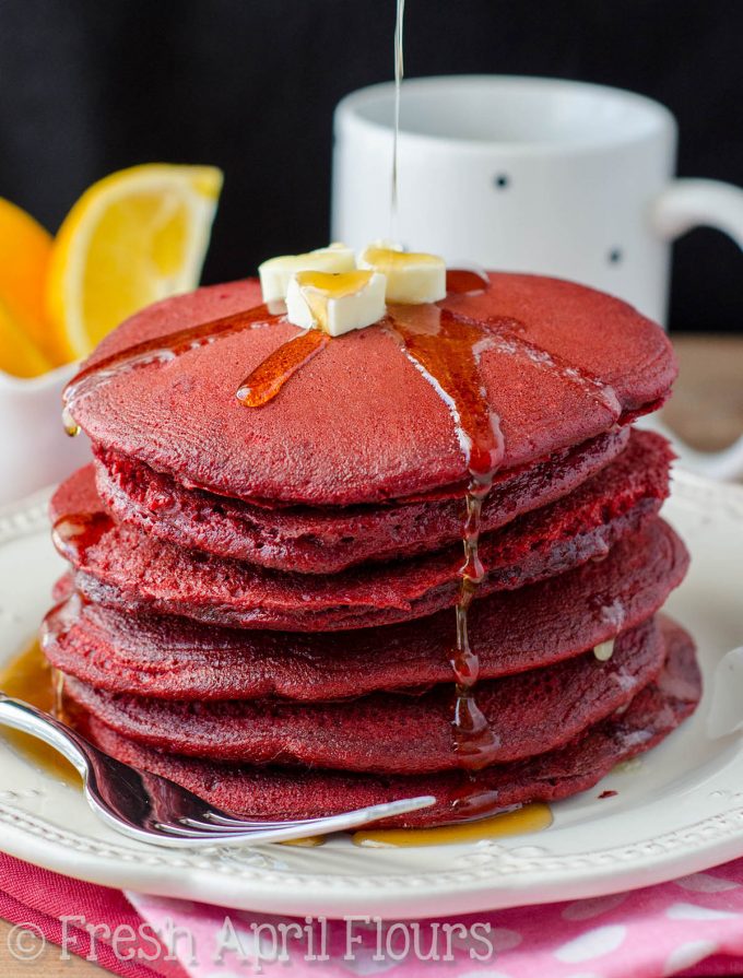 A stack of red velvet pancakes with syrup getting poured onto the top.