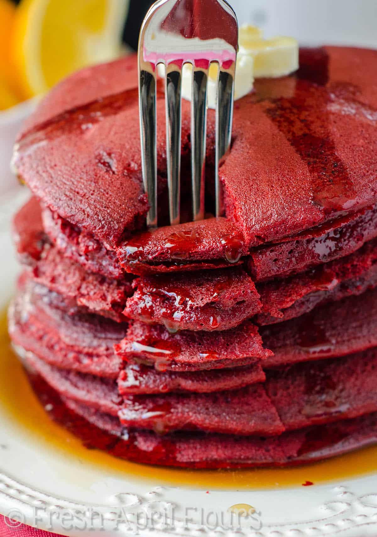 A fork digging into a stack of red velvet cake mix pancakes with syrup dripping down the stack.