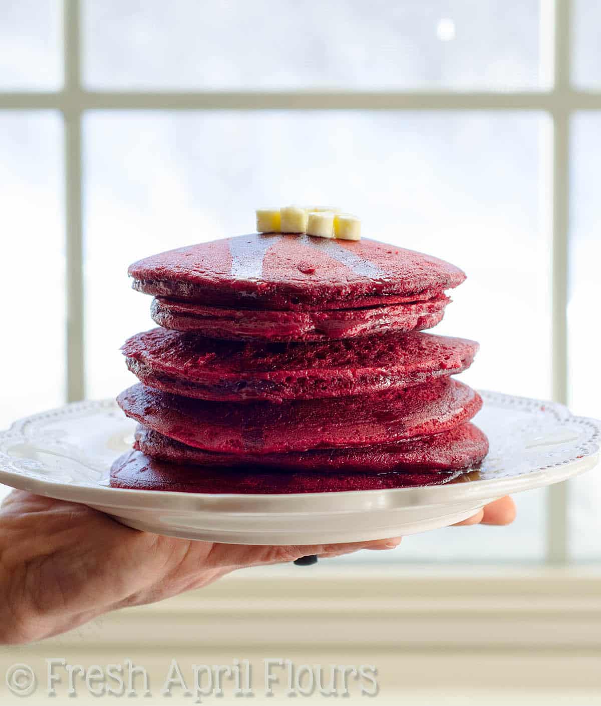 A hand holding a plate of a stack of red velvet pancakes.