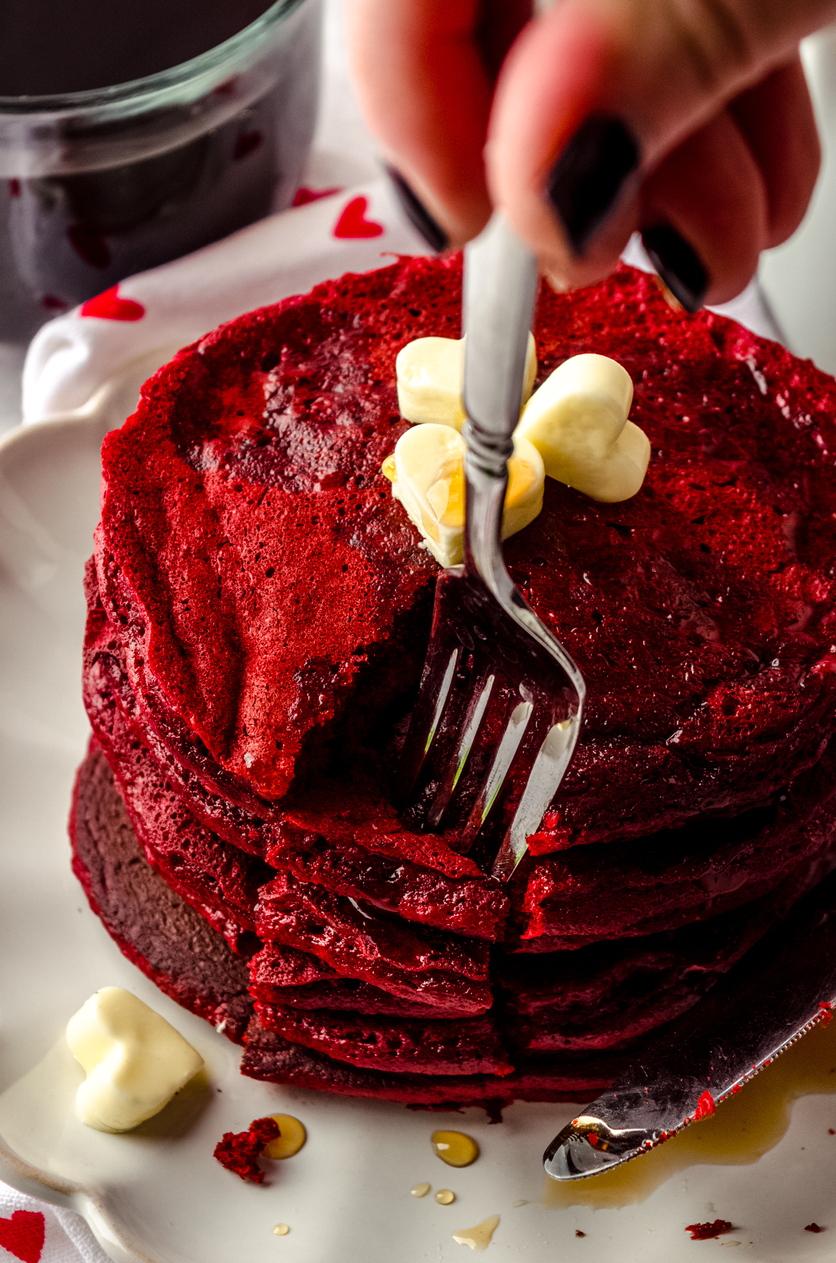 A fork digging into a stack of red velvet pancakes. There is syrup dripping down the sides and little butter hearts on top of the pancakes.