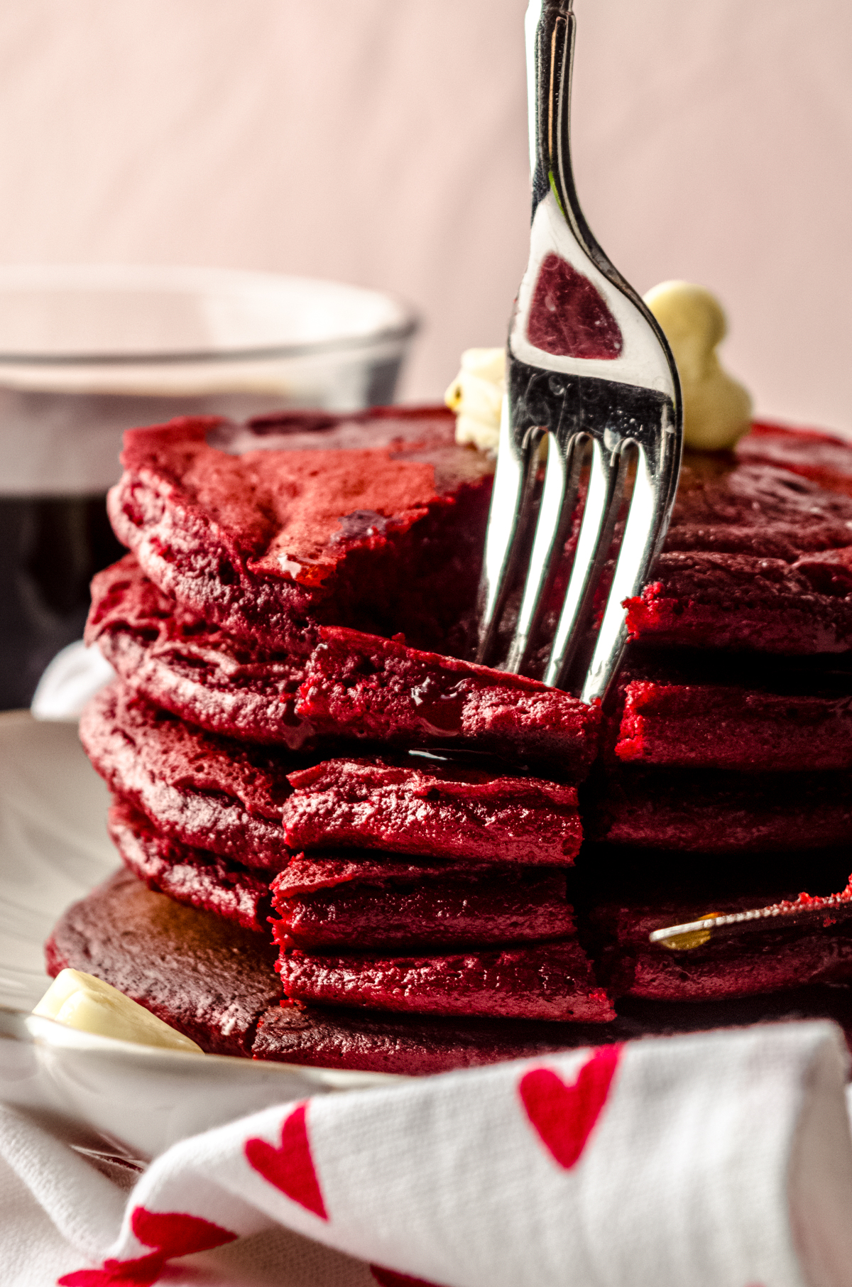 A fork digging into a stack of red velvet pancakes. There is syrup dripping down the sides and little butter hearts on top of the pancakes.