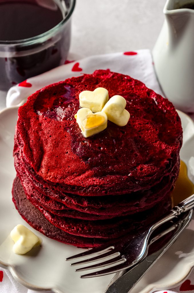 A stack of red velvet pancakes on a plate with a fork. There are mini butter hearts on top of the pancakes and a cup of coffee and syrup container in the background.