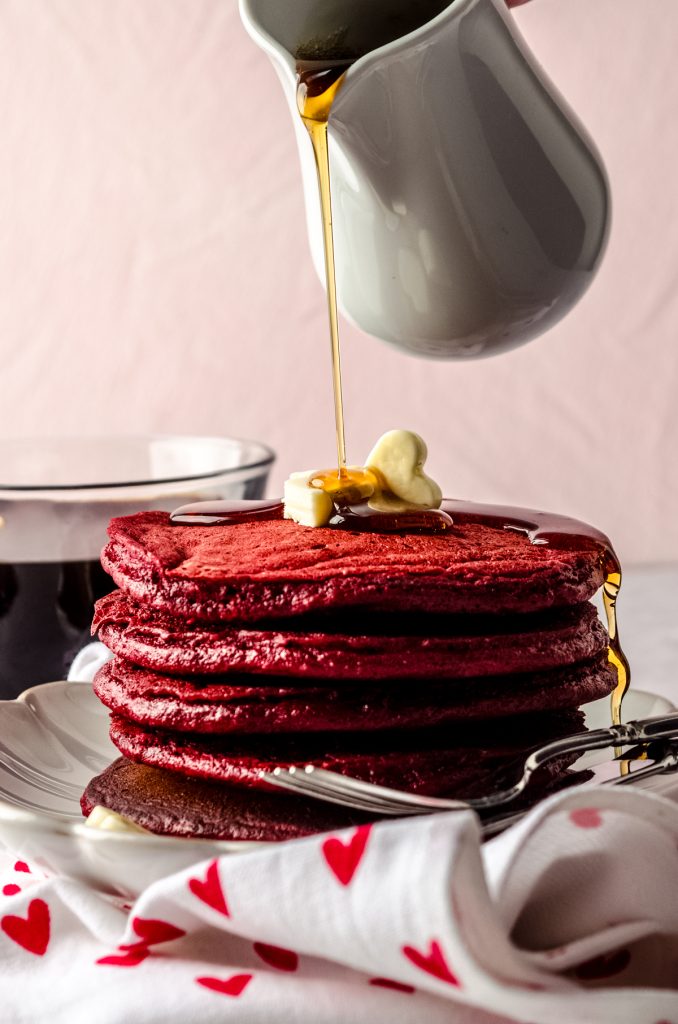Someone is pouring syrup onto a stack of red velvet pancakes.