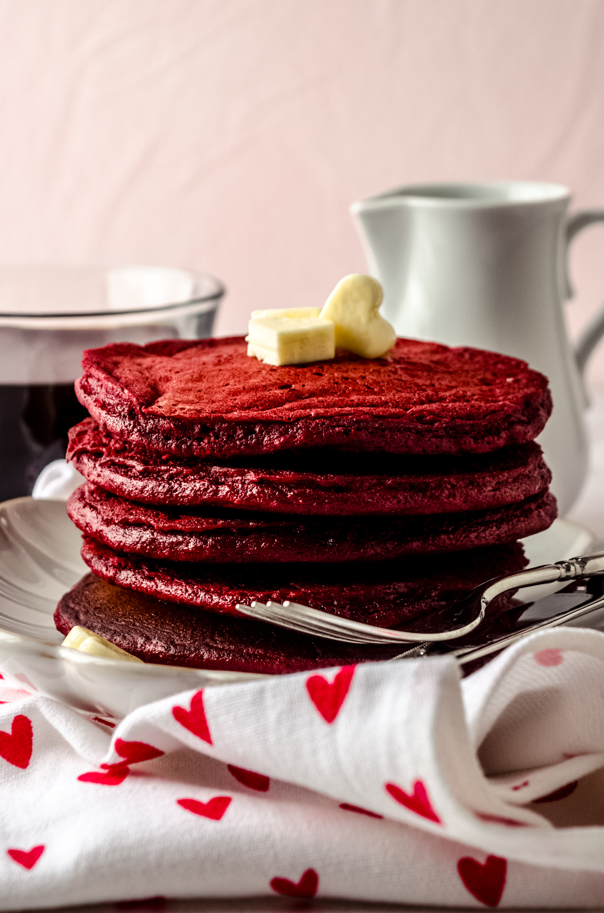 A stack of red velvet pancakes on a plate with a fork. There are mini butter hearts on top of the pancakes and a cup of coffee and syrup container in the background.