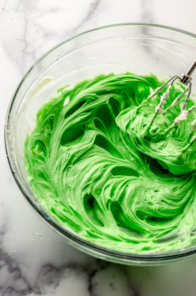 A bowl of beaten cream cheese mixed with green food coloring for no bake mint chocolate chip pie with beaters in it.