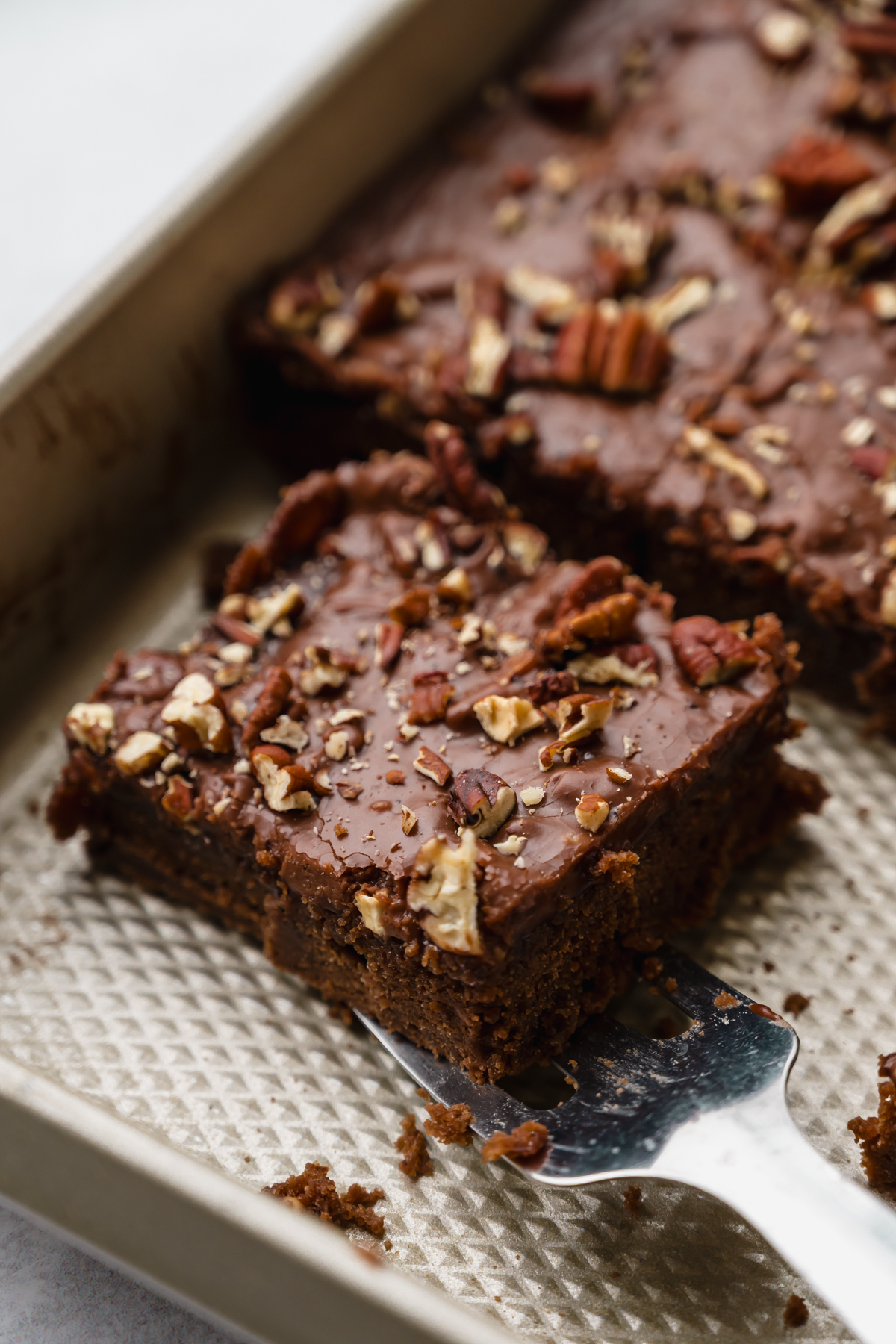 Someone is lifting a slice of chocolate Texas sheet cake out of a pan with a spatula.