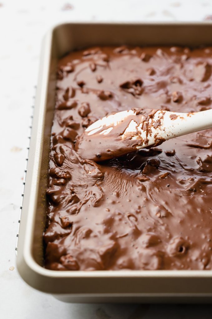 Someone is spreading chocolate icing onto a baked chocolate Texas sheet cake with a spatula.