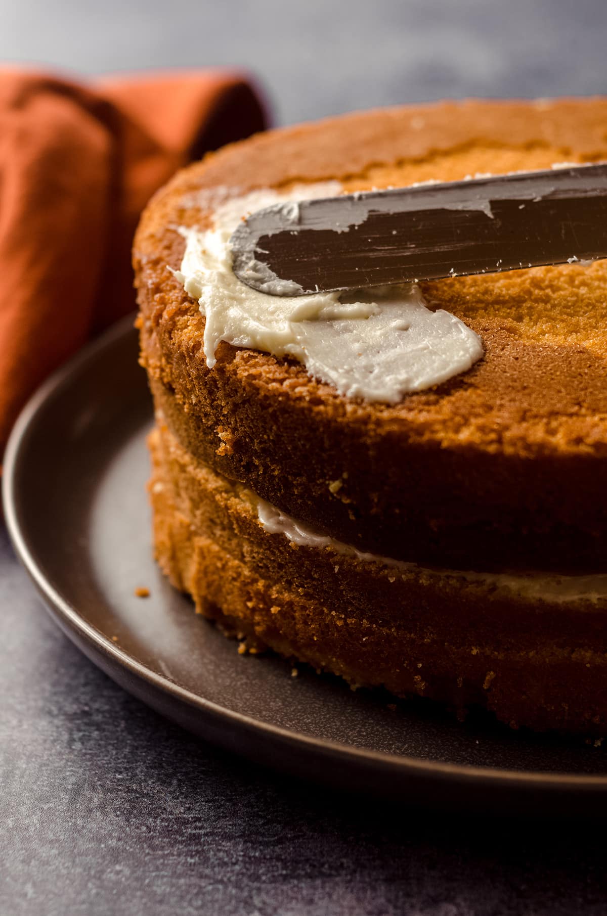 stacked orange creamsicle layer cake being covered in orange cream cheese frosting with a metal spatula