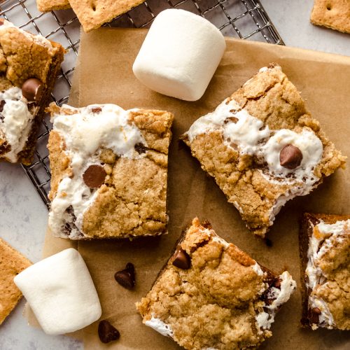 Aerial photo of s'mores bars on a surface with a wire rack, chocolate chips, and marshmallows.