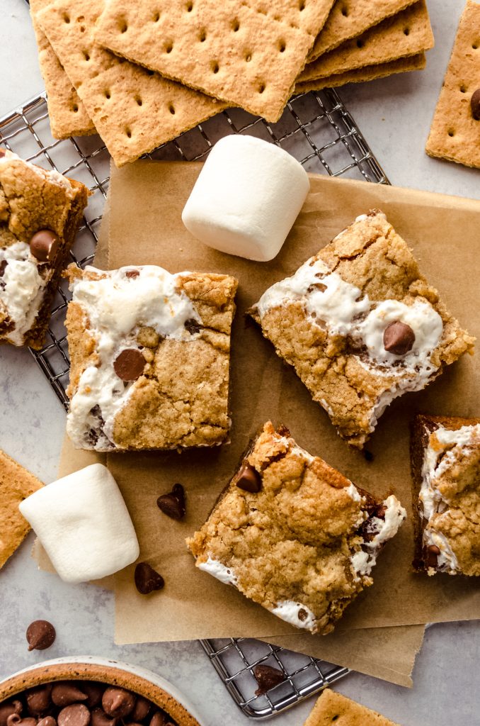 Aerial photo of s'mores bars on a surface with a wire rack, chocolate chips, and marshmallows.