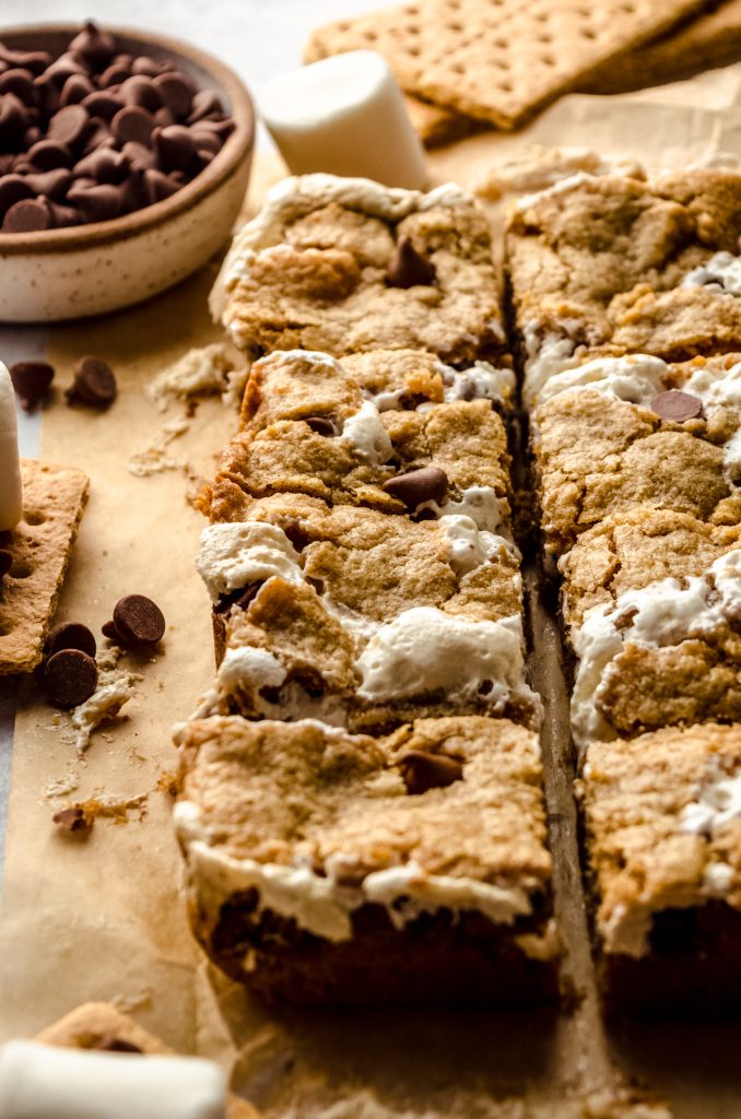 S'mores bars on parchment with chocolate chips, graham crackers, and marshmallows around it.