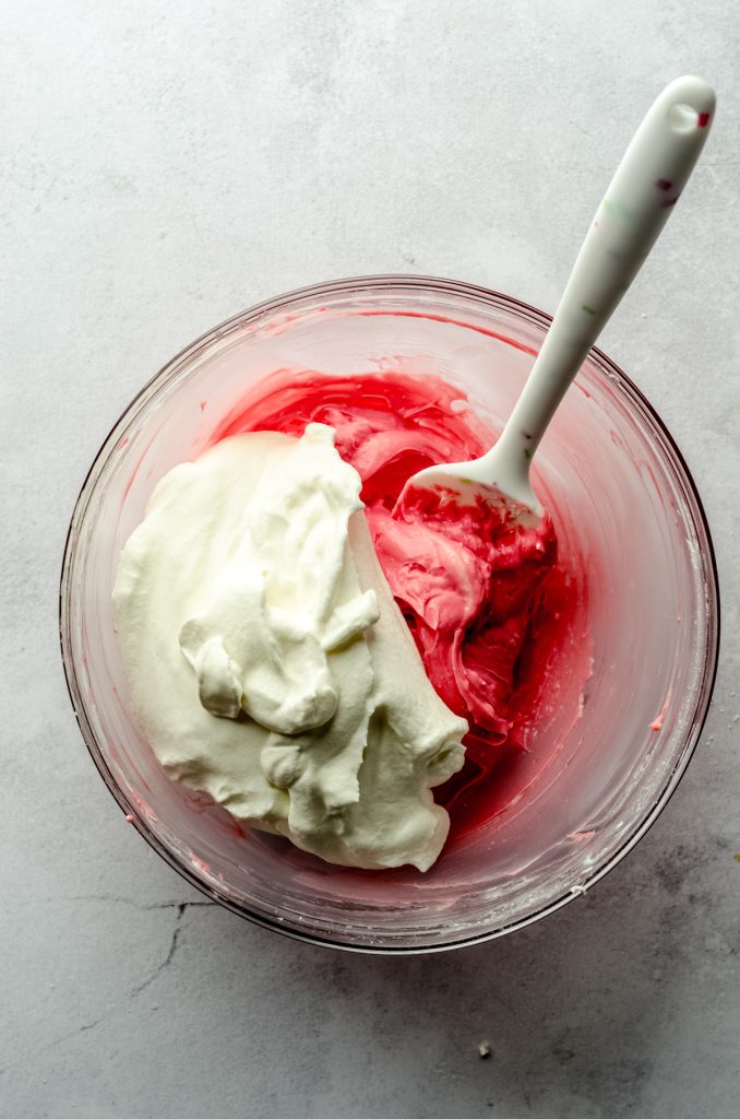 Aerial photo of a cream cheese and red food coloring filling with whipped cream dolloped into it and ready to stir with a spatula that's sitting in the bowl.