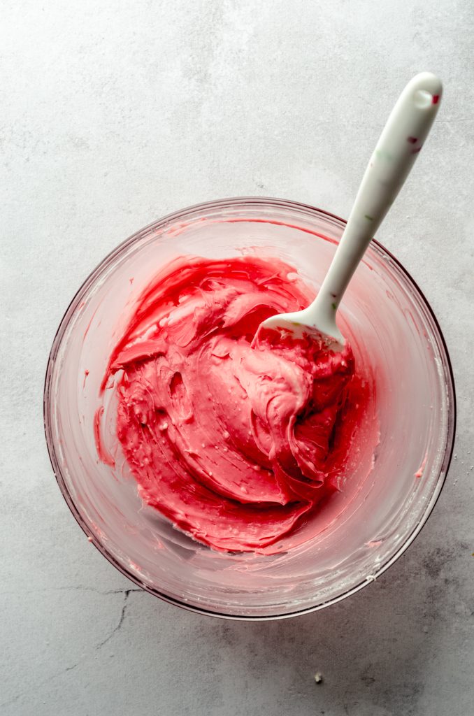Aerial photo of cream cheese and red food coloring with a spatula in it.