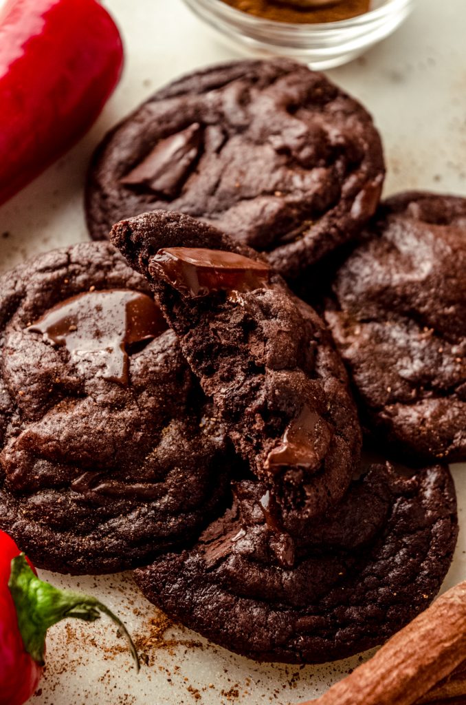 A chocolate cayenne cookie that has been split in half with a cinnamon stick, chili pepper, and nutmeg around it.