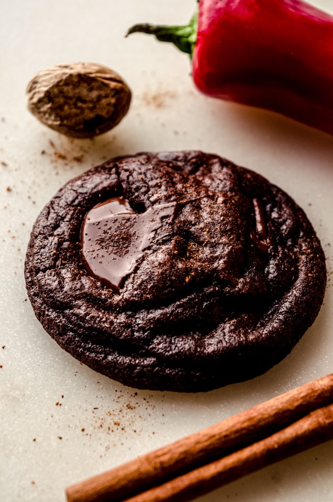 A chocolate cayenne cookie with a cinnamon stick, chili pepper, and nutmeg around it.