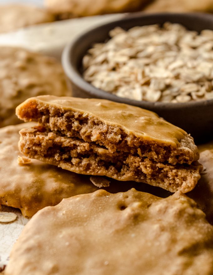 Molasses iced oatmeal cookies on a plate with a bowl of oats in the background and the cookie in the front has been broken in half.