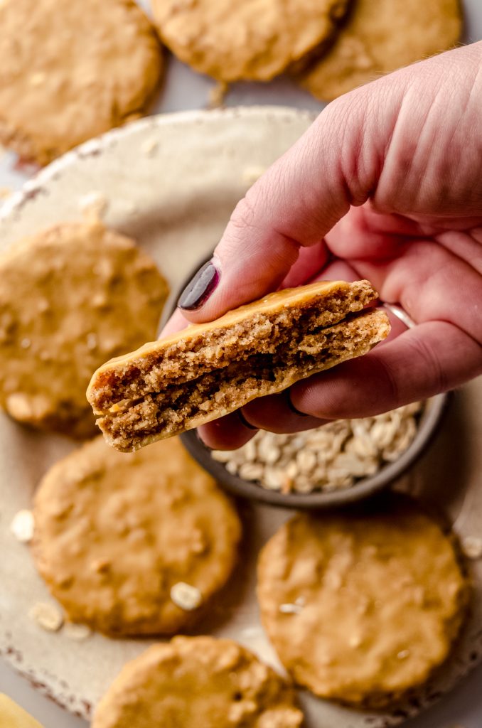 Aerial photo of someone holding a molasses iced oatmeal cookie above a a plate with a bowl of oats and the cookie has been broken in half.