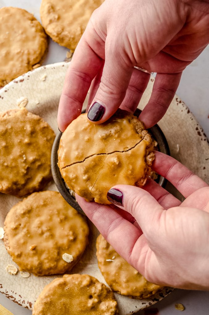 Aerial photo of someone holding a molasses iced oatmeal cookie above a a plate with a bowl of oats and breaking it in half.