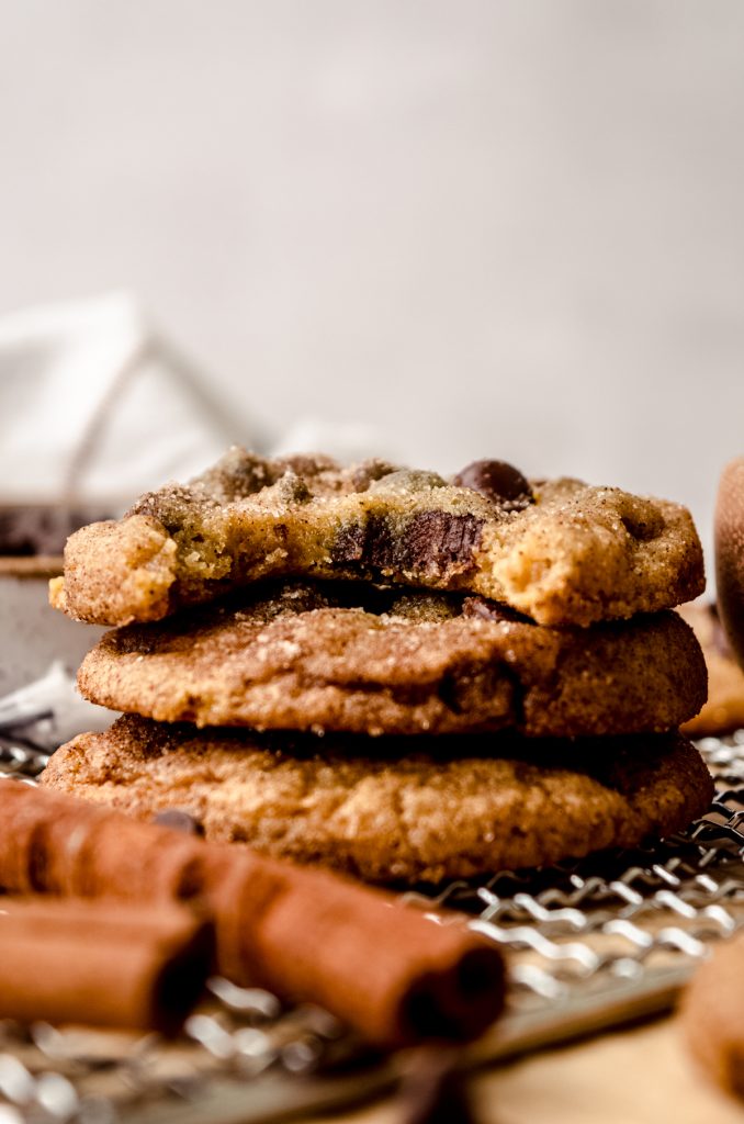 A stack of snickerdoodle chocolate chip cookies on a cooling rack with cinnamon sticks in the foreground and a bite taken out of the cookie on the top.