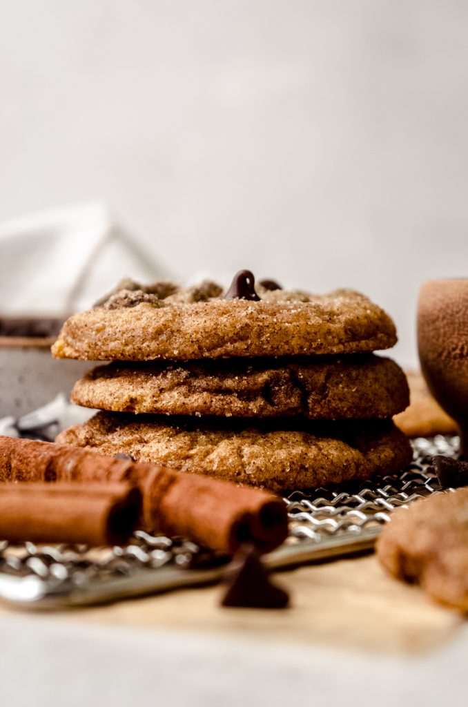 A stack of snickerdoodle chocolate chip cookies on a cooling rack with cinnamon sticks in the foreground.