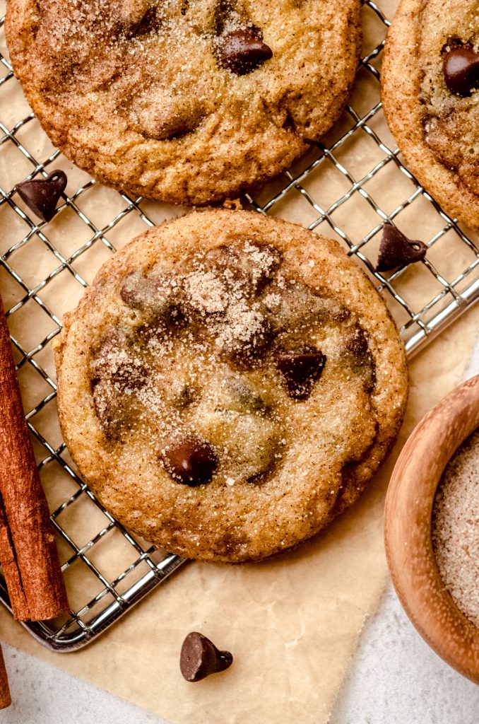 Aerial photo of chocolate chip snickerdoodle cookies on a cooling rack with cinnamon sticks.