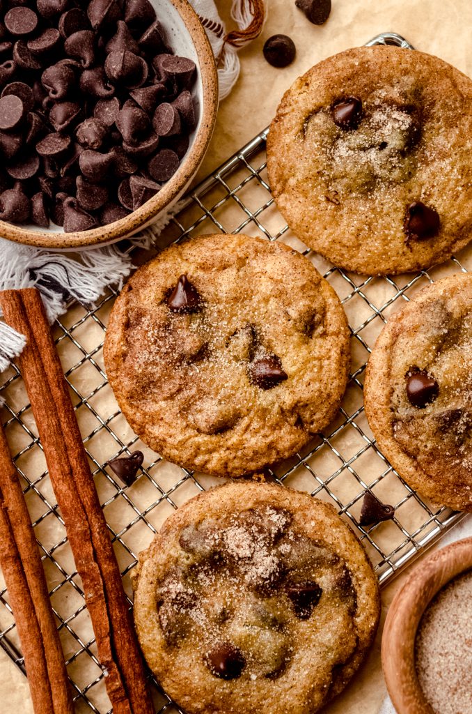 Aerial photo of chocolate chip snickerdoodle cookies on a cooling rack with cinnamon sticks and a bowl of chocolate chips.