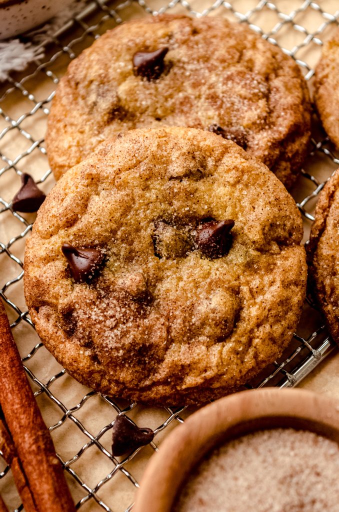 A chocolate chip snickerdoodle cookie on a cooling rack with cinnamon sticks and a bowl of cinnamon-sugar.