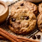 A chocolate chip snickerdoodle cookie on a cooling rack with cinnamon sticks and a bowl of cinnamon-sugar.