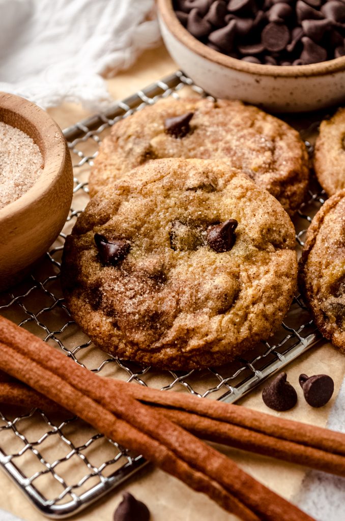 A chocolate chip snickerdoodle cookie on a cooling rack with cinnamon sticks and a bowl of cinnamon-sugar.