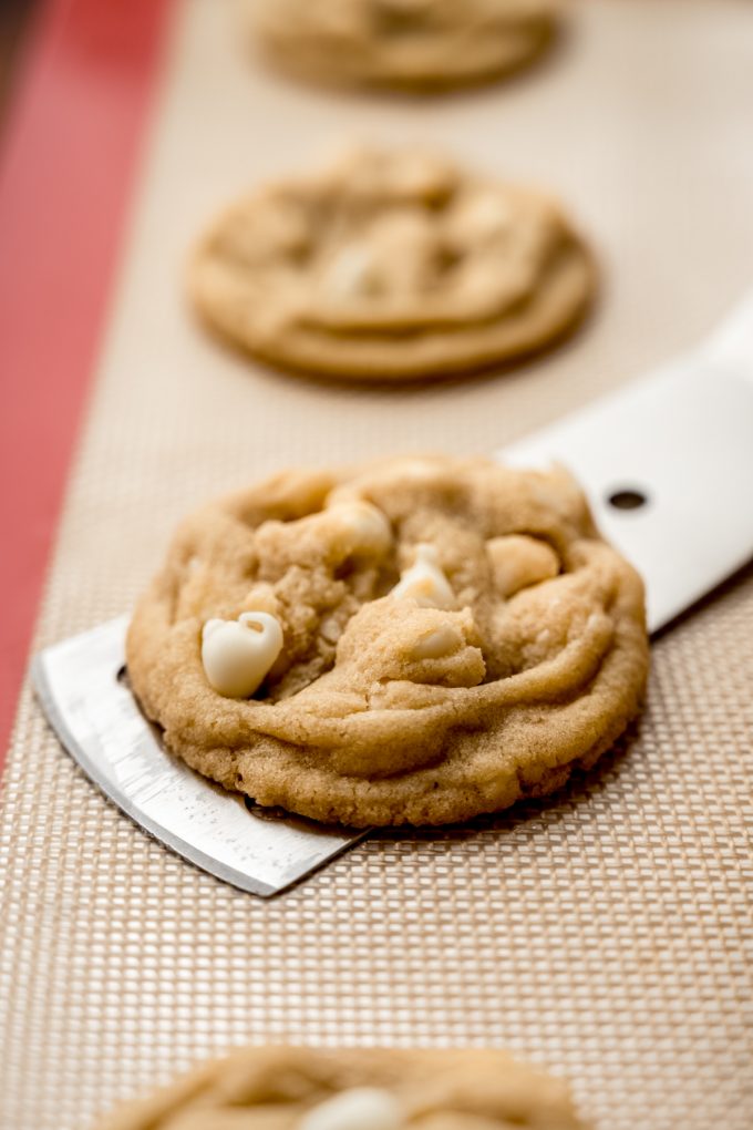 Someone is using a spatula to lift a white chocolate macadamia nut cookie off of a baking sheet.