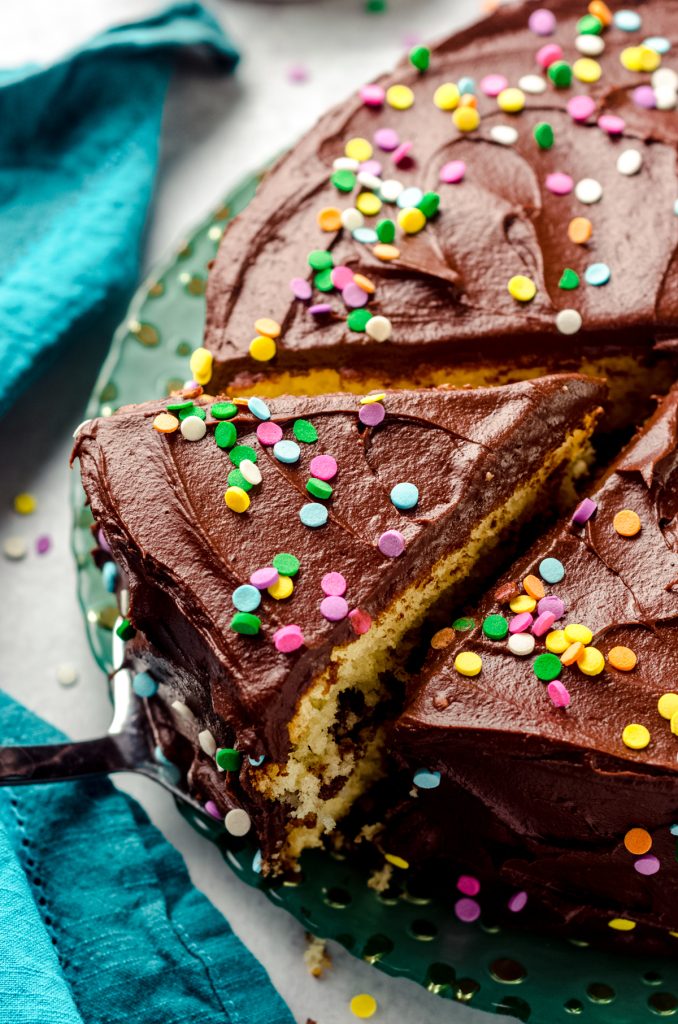 Yellow cake with chocolate frosting and rainbow confetti quins on a plate. Someone is removing a slice with a cake server.