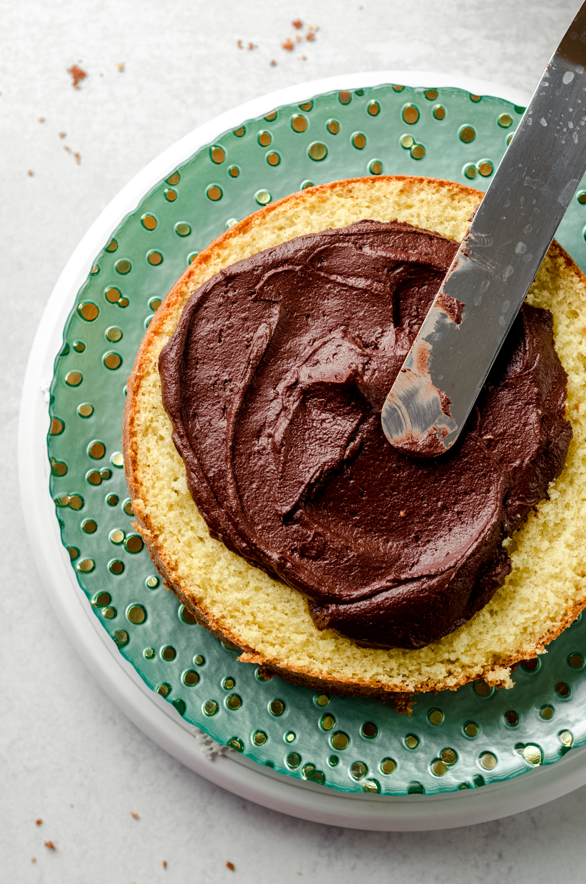 Aerial photo of someone using a spatula to spread chocolate frosting onto a layer of yellow cake.