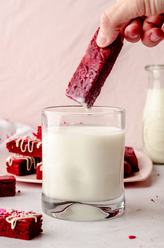 Someone dunking a red velvet biscotti into a glass of milk.