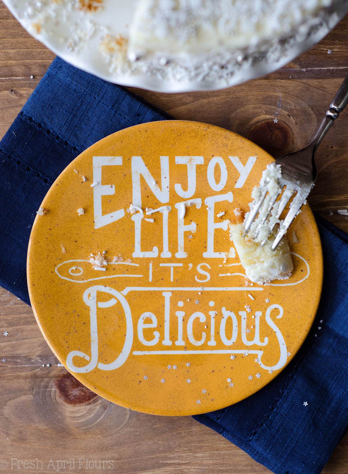 yellow plate that says "enjoy life it's delicious" with cake crumbs and a fork with one small bite of white layer cake on it