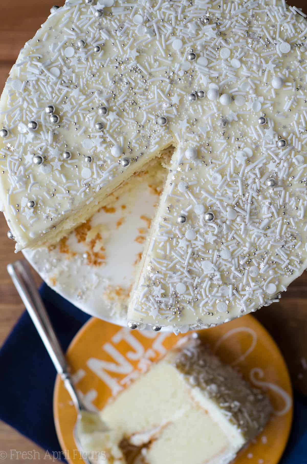 aerial photo of white layer cake with white and silver sprinkles all over it and a slice of cake removed and on a yellow plate in the background