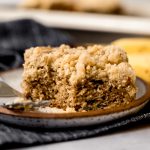 A slice of banana crumb cake on a plate with a fork and a bite taken out of the cake.