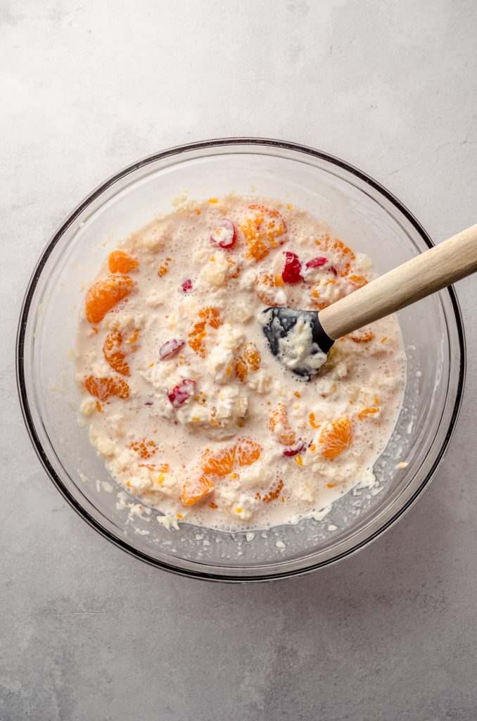 Aerial photo of canned pineapple, mandarin oranges, maraschino cherries, and sour cream in a bowl to make ambrosia salad.