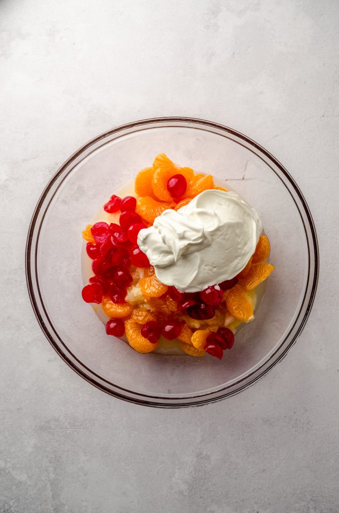 Aerial photo of canned pineapple, mandarin oranges, maraschino cherries, and sour cream in a bowl to make ambrosia salad.