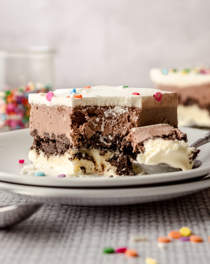 A slice of ice cream sheet cake on a plate with a spoon and a bite taken out of it.